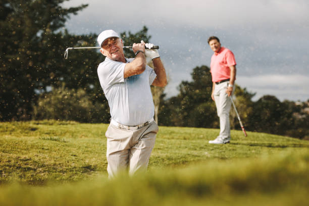 Pro golfer playing on the course with second player standing at back looking on. Man hitting the ball out of a sand bunker during the game.